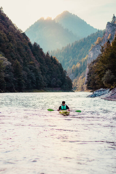 Young man kayaking on the Dunajec river, sitting in a kayak and paddling down the river. Enjoying ride surrounded by hills and beautiful view of valley and mountains peaks. Spends vacation on wandering with backpacks in a mountains and forests