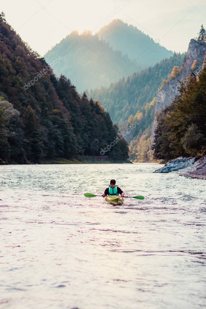 Young man kayaking on the Dunajec river, sitting in a kayak and paddling down the river. Enjoying ride surrounded by hills and beautiful view of valley and mountains peaks. Spends vacation on wandering with backpacks in a mountains and forests