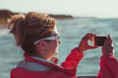 Genç kadının telefonuyla, fotoğraflar çekmek seyir perde açık havada duran, o düz gökyüzü ve denizin içinde belgili tanımlık geçmiş ile güneş ışığı tarafından backlighted