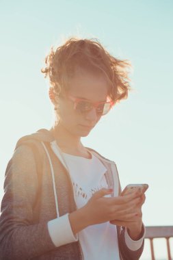 Seyir perde telefon, kullanan genç sarışın kız açık havada duran, o arka planda düz gökyüzü güneş ışığı tarafından backlighted