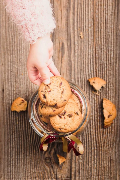 Little girl taking a cookie from a jar filled with oat cookies standing on a wooden table