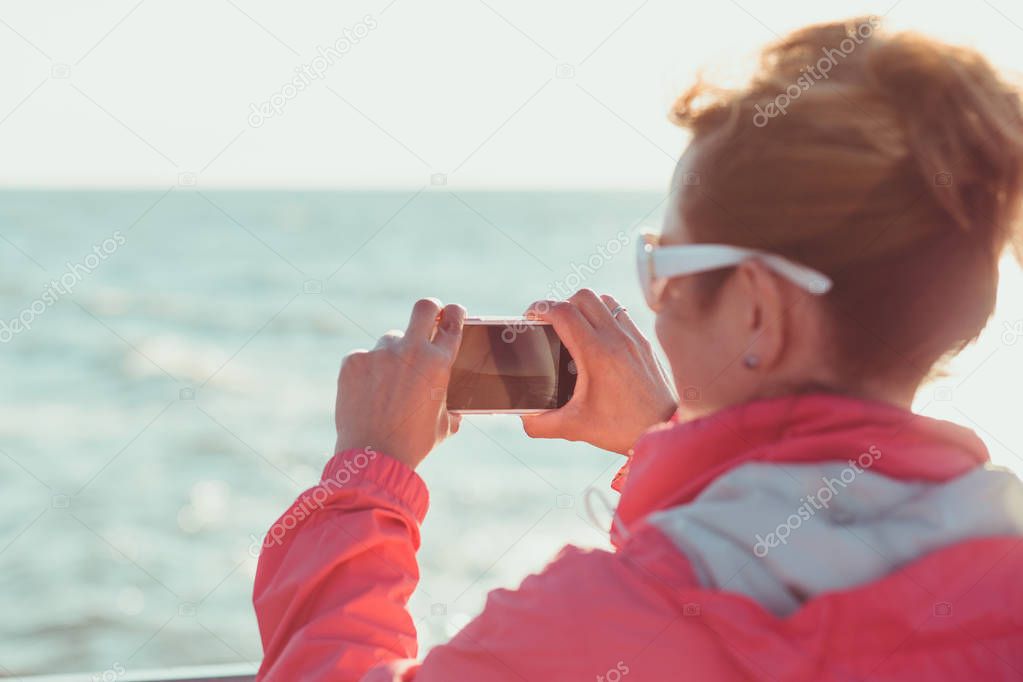 Young woman taking a photos using phone, looking at screen, standing outdoors, she is backlighted by sunlight with plain sky and sea in the background