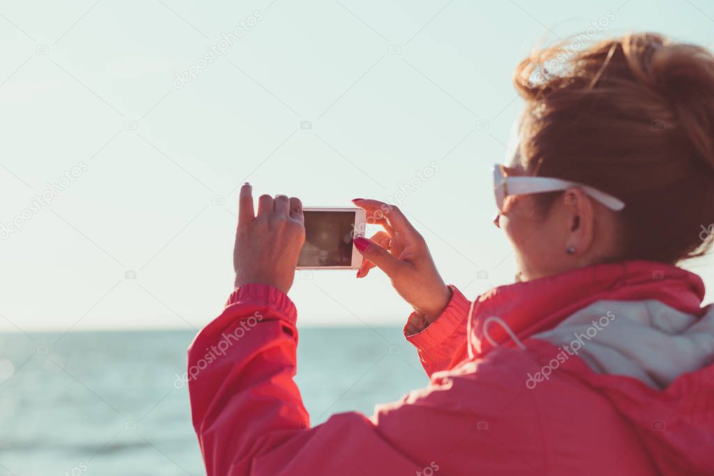 Young woman taking a photos using phone, looking at screen, standing outdoors, she is backlighted by sunlight with plain sky and sea in the background