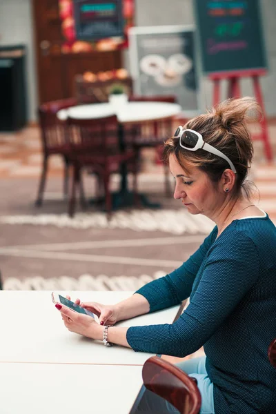 Młoda kobieta za pomocą telefonu komórkowego siedzi przy stole w kawiarni — Zdjęcie stockowe
