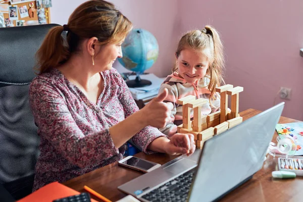 Woman mother working doing her job remotely during video chat call stream online course webinar on laptop from home while her daughter playing with bricks toy. Woman sitting at desk in front of computer looking at screen