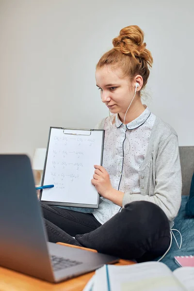 Young woman student having classes, learning online, watching lesson remotely, listening to professor, talking with classmates on video call from home during quarantine. Young girl using laptop, headphones, books, manuals sitting on bed