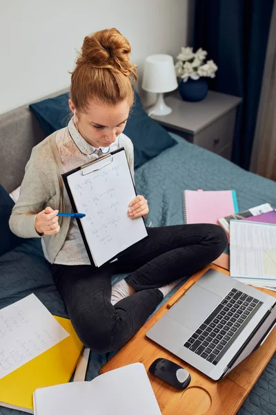 Young woman student having classes, learning online, watching lesson remotely, listening to professor, talking with classmates on video call from home during quarantine. Young girl using laptop, headphones, books, manuals sitting on bed