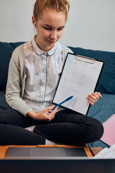 Young woman student having classes, learning online, watching lesson remotely, listening to professor, talking with classmates on video call from home during quarantine. Young girl using laptop, headphones, books, manuals sitting on bed