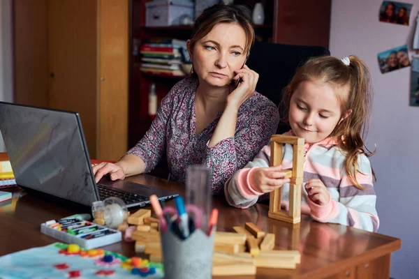 Woman mother working doing her job remotely during video chat call stream online course webinar on laptop from home while her daughter playing with bricks toy. Woman sitting at desk in front of computer looking at screen