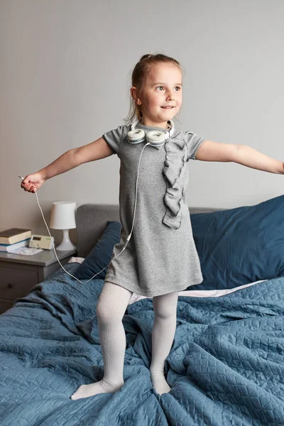 小女孩拿着耳机唱歌 模仿自己是个真正的歌手 在家里的卧室里 孩子们在床上听音乐 跳起舞来很开心 — 图库照片