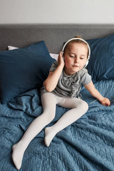 Little girl singing holding headphones cord imitating herself a real singer. Child having fun jumping dancing listening to music on bed in bedroom at home
