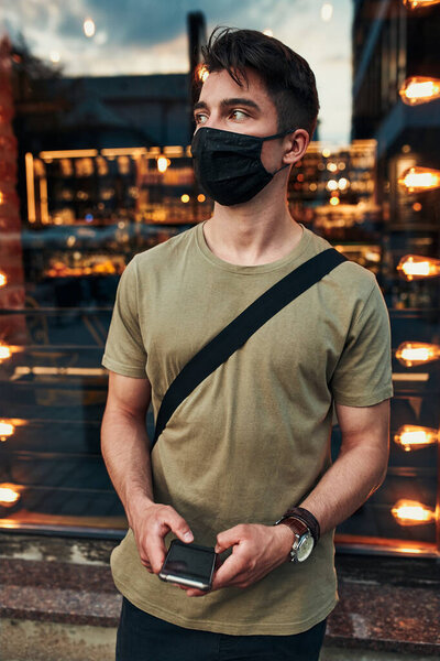 Young man standing at store front in the city center in the evening, looking away, holding smartphone, wearing the face mask to avoid virus infection and to prevent the spread of disease in time of coronavirus