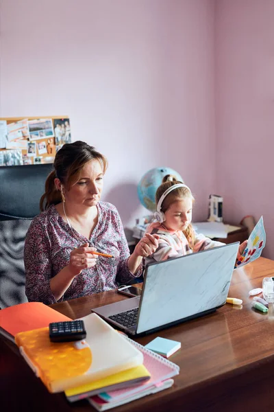 Woman mother working doing her job remotely during video chat call stream online course webinar on laptop from home while her daughter making drawings playing around and listening to stories. Woman sitting at desk in front of computer looking at scre