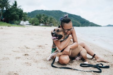 Cheerful pretty young woman in eyeglases sitting and hugging her dog on the beach clipart