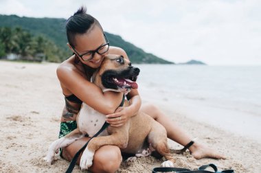 Cheerful pretty young woman in eyeglases sitting and hugging her dog on the beach clipart