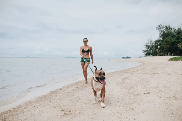 ビーチで彼女の犬を連れて歩いて陽気な若い女性 — ストック写真