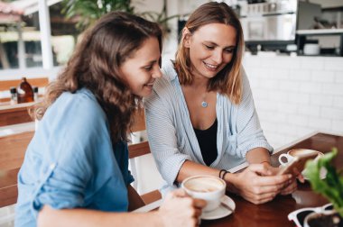 Cheerful female friends chatting in cafe. Two beautiful happy women looking in smartphone. and drinking coffee. Friendship concept