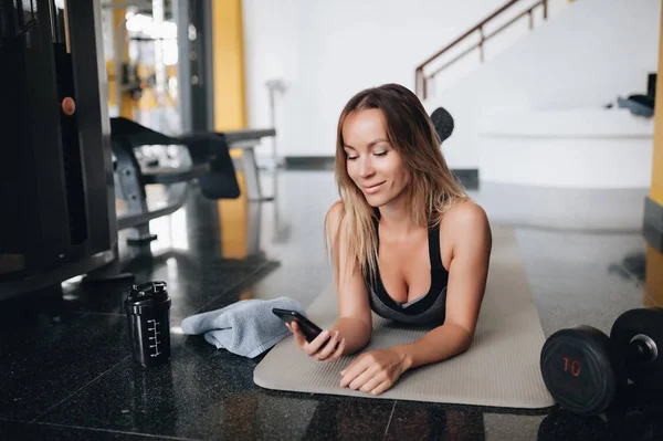 Atractiva mujer caucásica en ropa deportiva está utilizando un teléfono inteligente y sonriendo mientras se pone en la esterilla de yoga —  Fotos de Stock