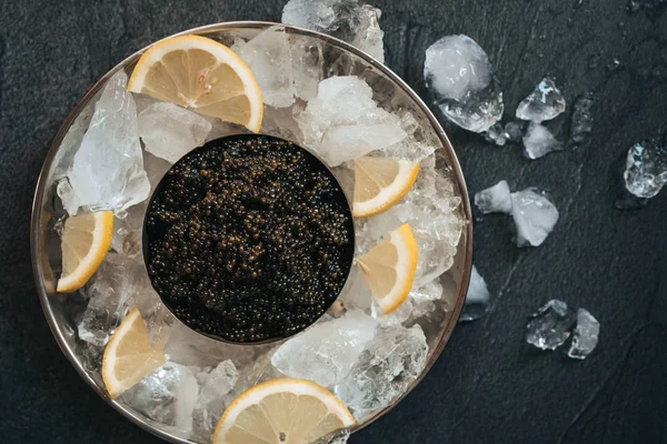 Μαύρο χαβιάρι στο πιάτο με πάγο και λεμόνι στο μαύρο τραπέζι — Φωτογραφία Αρχείου