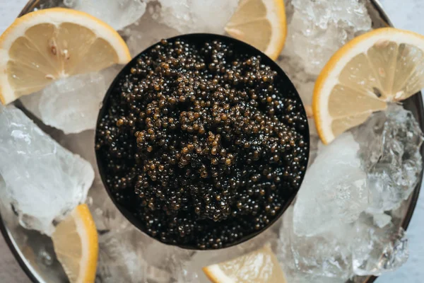 Черная икра осетра в банке со льдом и лимоном на белом фоне — стоковое фото