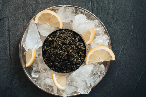 Μαύρο χαβιάρι στο πιάτο με πάγο και λεμόνι στο μαύρο τραπέζι — Φωτογραφία Αρχείου