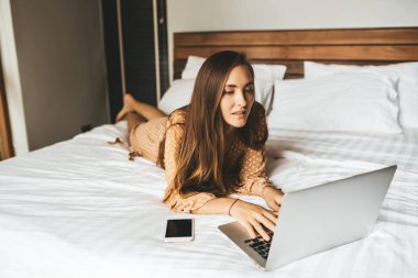 Uzun saçlı, bacak bacak üstüne atmış ve laptopla yatak odasında çalışan bir kadın.