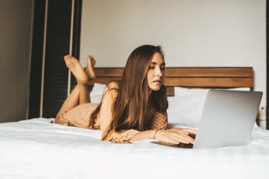Uzun saçlı, bacak bacak üstüne atmış ve laptopla yatak odasında çalışan bir kadın.