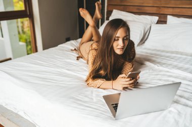 Uzun saçlı kadın yatak odasında dizüstü bilgisayarla telefonda sohbet ediyor.