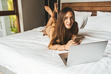 Uzun saçlı kadın yatak odasında dizüstü bilgisayarla telefonda sohbet ediyor.