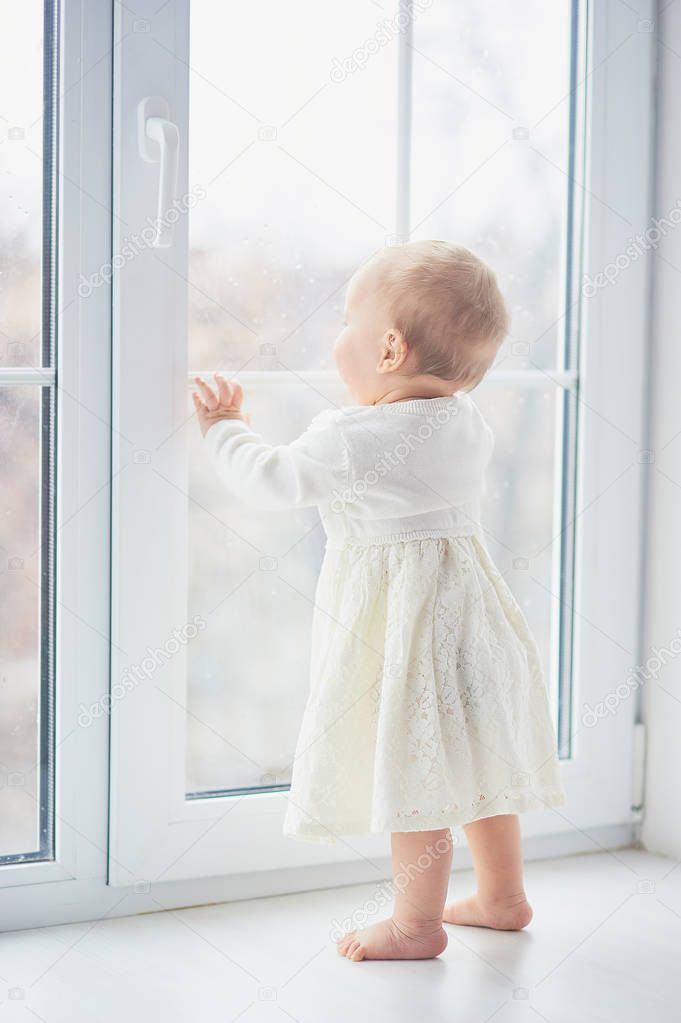 Blonde curly toddler Baby girl looking through a window. Toddler is dreaming, looking at the street outside. safe window.