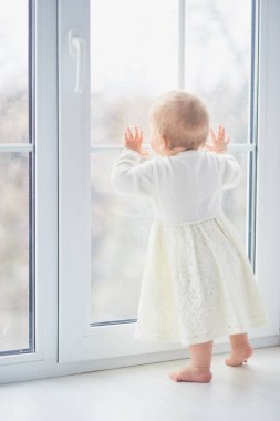 Sarışın kıvırcık toddler bebek kız bir pencereden bakıyor. Yürümeye başlayan çocuk rüya görüyor, sokağın seyir dışında. güvenli pencere.
