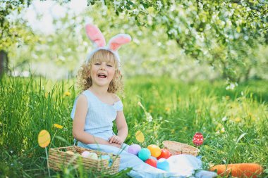 Paskalya yumurtaları ve bahçesinde tavşan kulakları olan sevimli, komik bir kız. Paskalya konsepti. Paskalya yumurtası avında gülen çocuk. Sepet dolusu yumurta ve bahar konseptiyle parkta bir çocuk.
