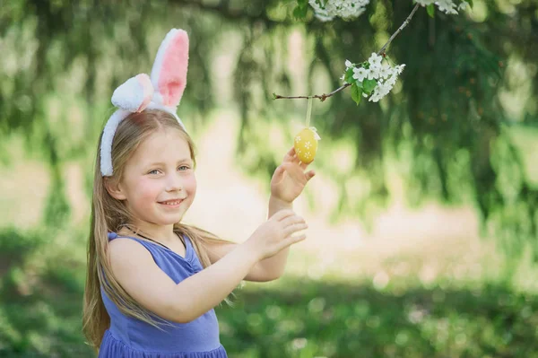 Милая забавная девушка с пасхальными яйцами и кроличьими ушами в саду. Пасхальная концепция. Смеющийся ребенок во время охоты за пасхальными яйцами . — стоковое фото