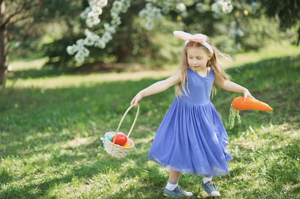Милая забавная девушка с пасхальными яйцами и кроличьими ушами в саду. Пасхальная концепция. Смеющийся ребенок во время охоты за пасхальными яйцами . — стоковое фото