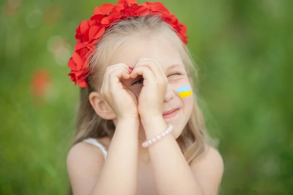 Kleines ukrainisches Mädchen, das durch eine Herzgeste blickt, die mit Händen im sommergrünen Park gemacht wurde. Geste der Liebe zur Ukraine durch ein hübsches kleines Kind im Mohnfeld. — Stockfoto