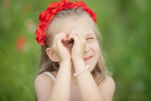 Pequena menina ucraniana olhando através do gesto do coração feito com as mãos no verão parque verde. Gesto de amor para a Ucrânia por criança muito jovem no campo de papoula . — Fotografia de Stock