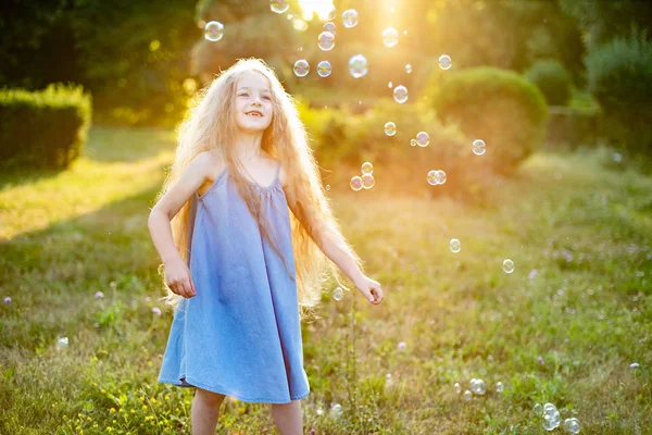 Enfant tourbillonnant, dansant joue sur la prairie. Fille s'amuser avec des bulles. Jolie petite blonde à cheveux longs dansant avec des bulles de savon au parc du coucher du soleil — Photo