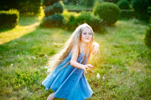 Дитячий свист, танці грають на лузі. Дівчина розважається з бульбашками. Симпатична маленька довгошерста блондинка танцює з мильними бульбашками в парку заходу сонця — стокове фото