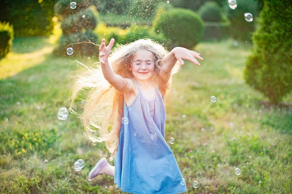 Παιδί που στροβιλίζεται, χορεύει στο λιβάδι. Το κορίτσι διασκεδάζει με τις φυσαλίδες. Χαριτωμένο μικρό ξανθό κορίτσι μακριά μαλλιά χορεύουν με σαπουνόφουσκες στο πάρκο ηλιοβασίλεμα — Φωτογραφία Αρχείου