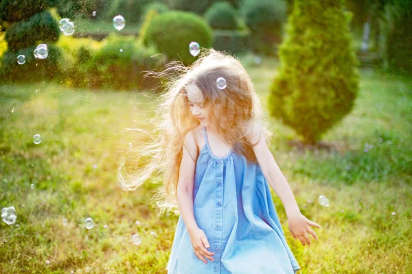 Dítě víří, tančí na louce. Holka se baví s bublinkama. Roztomilý malý dlouhosrstý blondýnka dívka tanec s mýdlem bubliny v parku západ slunce — Stock fotografie