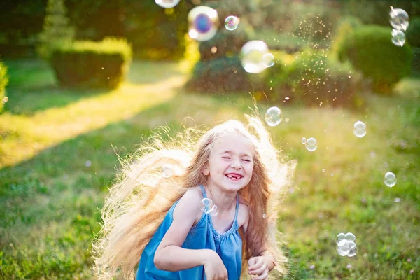 Дети кружатся, танцуют на лугу. Девочка развлекается с пузырьками. Милая блондинка с длинными волосами танцует с мыльными пузырями в парке заката — стоковое фото