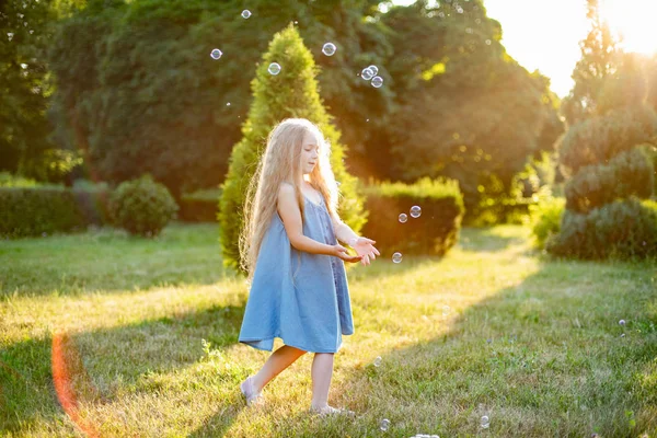 Un niño girando, bailando en el prado. Chica divirtiéndose con burbujas. Linda chica rubia de pelo largo bailando con burbujas de jabón en el parque del atardecer — Foto de Stock