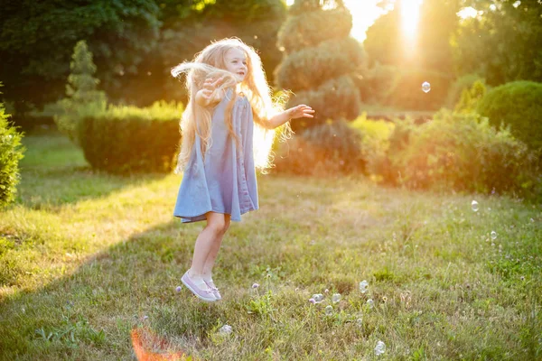 Anak berputar, menari bermain di padang rumput. Gadis bersenang-senang dengan gelembung. Gadis pirang berambut panjang yang lucu menari dengan gelembung sabun di taman matahari terbenam — Stok Foto