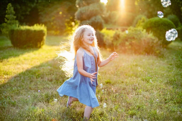 Enfant tourbillonnant, dansant joue sur la prairie. Fille s'amuser avec des bulles. Jolie petite blonde à cheveux longs dansant avec des bulles de savon au parc du coucher du soleil — Photo