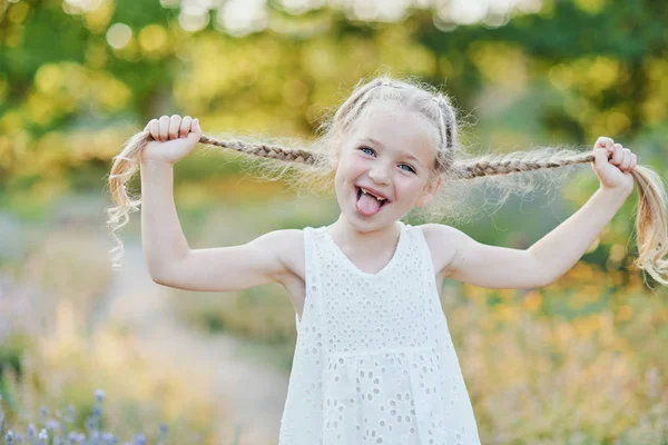 Funny girl holding hair plaits. Small cute girl with long blonde hair showing tongue outdoor. Kid expressing emotions. April fool. — Stock Photo, Image