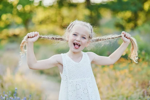 Rolig tjej som håller hårflätor. Liten söt tjej med långt blont hår som visar tungan utomhus. Kid uttrycker känslor. April Fool. — Stockfoto