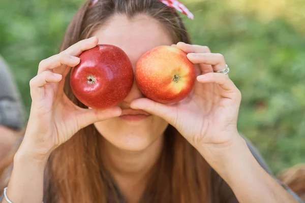 Girl holding apples in front of eyes while standing in orchard. Harvest concept