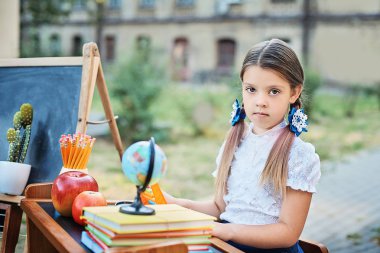 Portrait of a beautiful young first-grader sitting at a desk on the background autumn park. Farewell Bell. day of knowledge. beginning of the school year. school equipment pencils books, glass desk. clipart