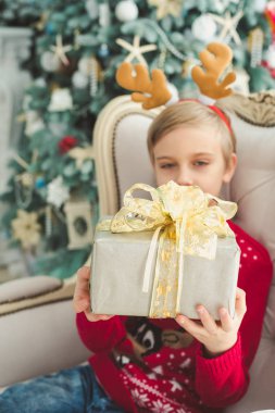 arka planda noel ağacı ile dekoratif odada koltukta otururken hediye kutusu tutan küçük çocuk