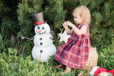 elf kostümü içinde küçük kız dekoratif yıldız tutan ve bahçede noel ağacı ve kardan adam yakınında oturan , juli noel kavramı
